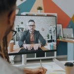 Equipe participa de uma videoconferência em um escritório colorido, com um colega exibido na tela do computador.