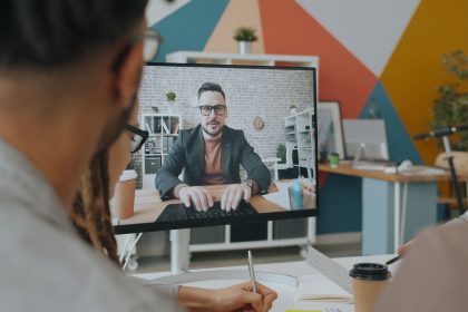 Equipe participa de uma videoconferência em um escritório colorido, com um colega exibido na tela do computador.
