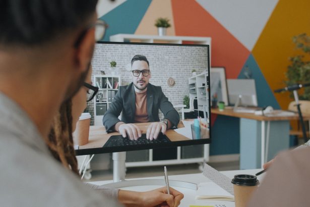 Equipe participa de uma videoconferência em um escritório colorido, com um colega exibido na tela do computador.