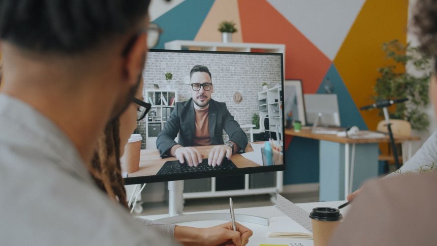 Equipe participa de uma videoconferência em um escritório colorido, com um colega exibido na tela do computador.
