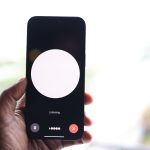 Mão segurando um smartphone com uma interface de assistente de voz exibida na tela.
