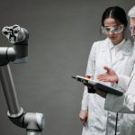 Pesquisadores usando jalecos e óculos de proteção analisam um braço robótico em um laboratório.