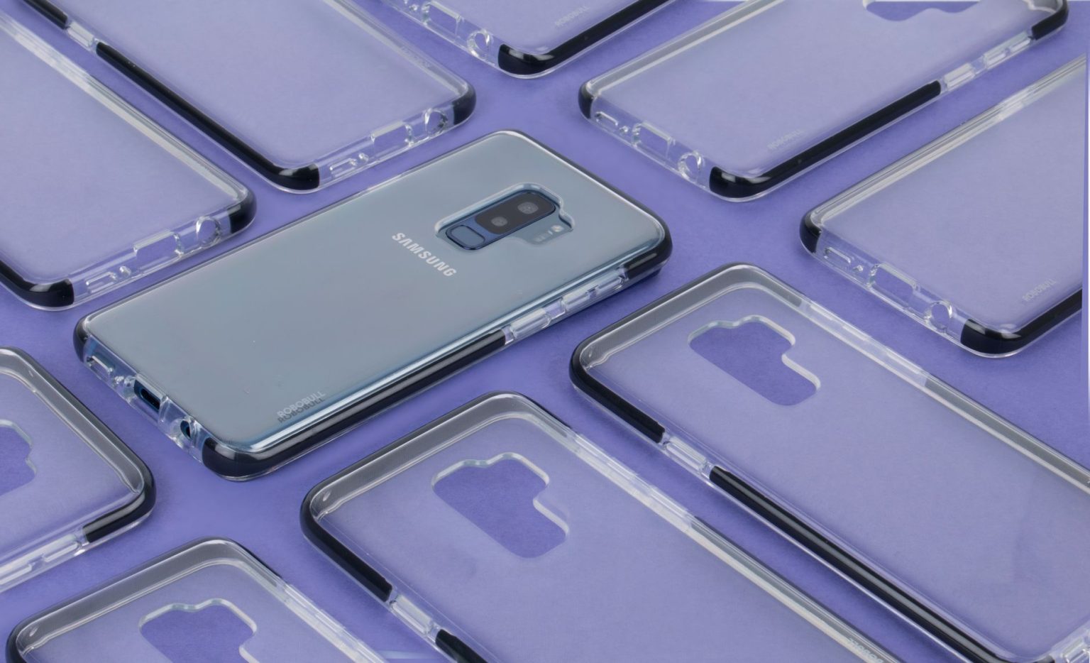 Close-up de um smartphone Samsung cercado por capas transparentes sobre uma superfície roxa.