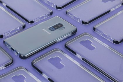 Close-up de um smartphone Samsung cercado por capas transparentes sobre uma superfície roxa.