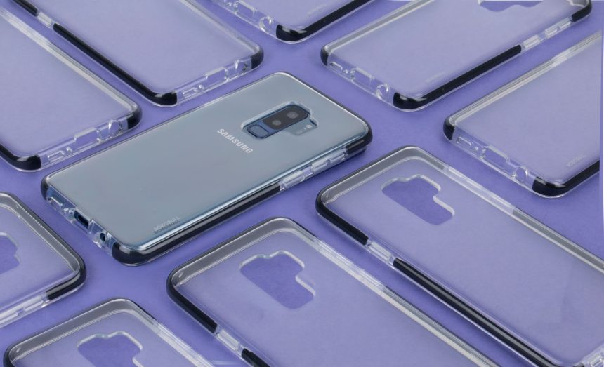 Close-up de um smartphone Samsung cercado por capas transparentes sobre uma superfície roxa.
