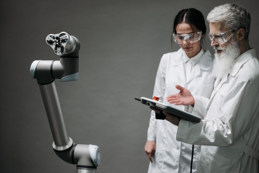 Pesquisadores usando jalecos e óculos de proteção analisam um braço robótico em um laboratório.
