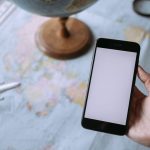 Mão segurando um smartphone sobre um mapa-múndi, ao lado de um globo e de um avião de brinquedo, simbolizando o planejamento de viagens.