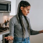 Mulher negra com tranças, em uma cozinha, gesticula durante uma conversa desagradável por videochamada no smartphone.
