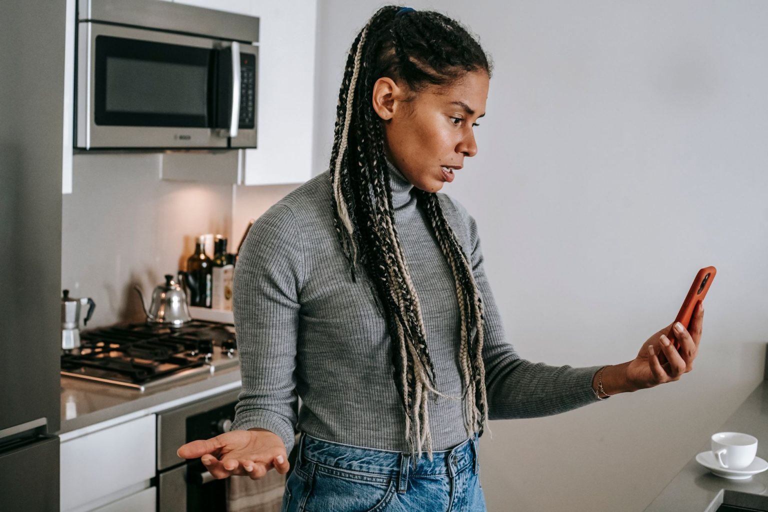 Mulher negra com tranças, em uma cozinha, gesticula durante uma conversa desagradável por videochamada no smartphone.