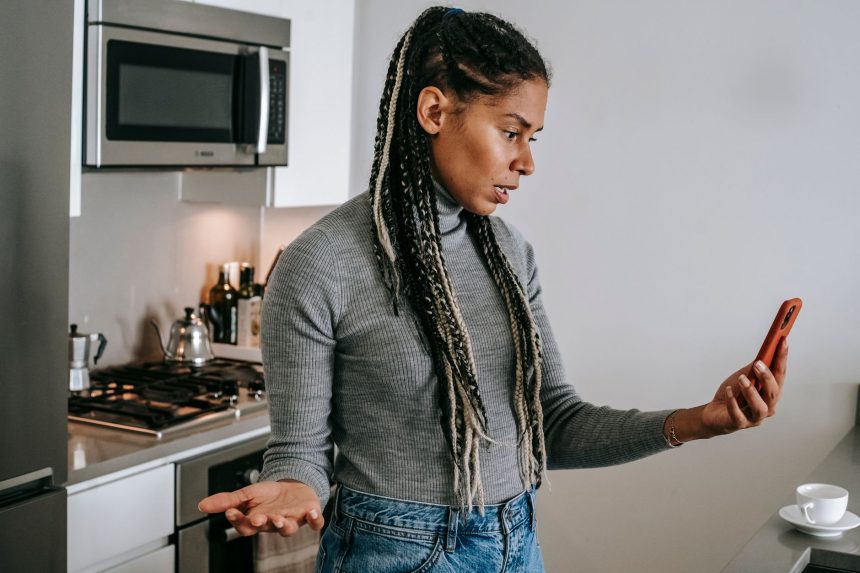 Mulher negra com tranças, em uma cozinha, gesticula durante uma conversa desagradável por videochamada no smartphone.